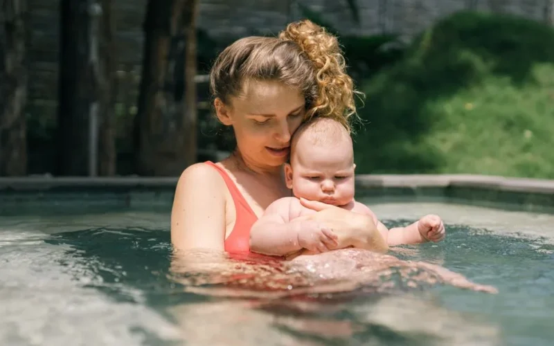 A loving mother holds her baby in a swimming pool, sharing a tender outdoors moment.