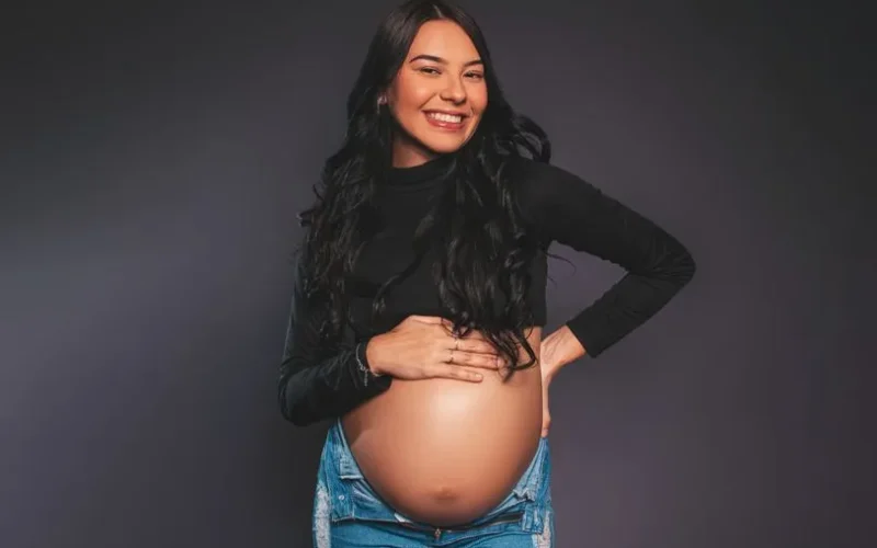 Confident pregnant woman posing and smiling against gray studio background.