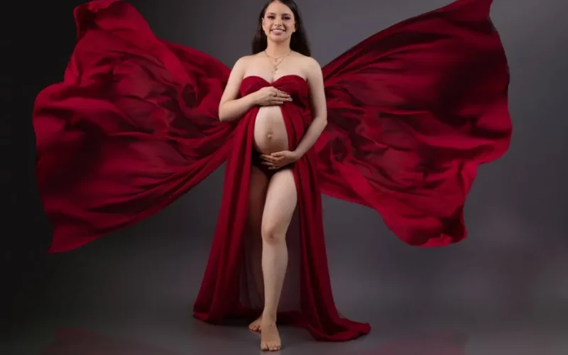 Studio portrait of a smiling pregnant woman in a flowing red gown, embracing motherhood.