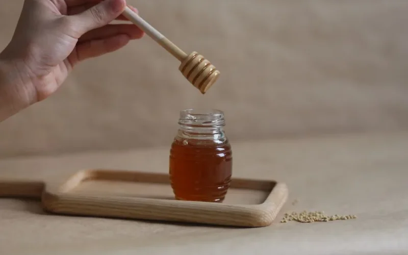 A hand holding a honey dipper over a jar on a wooden tray, showcasing the golden liquid.