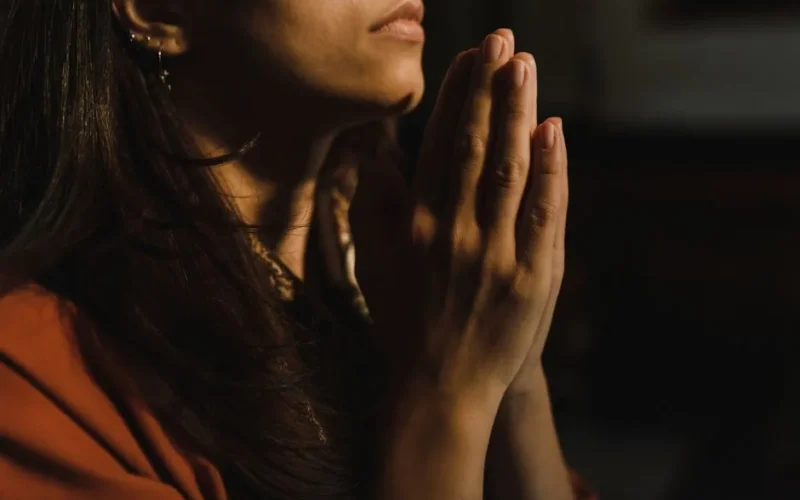 Close-up of a woman with folded hands praying, captured in a serene indoor setting with low lighting.