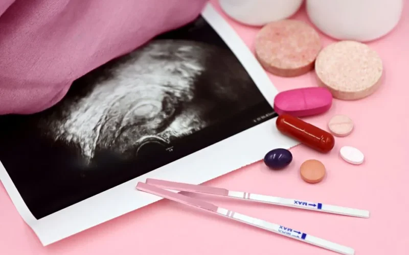 A closeup of pregnancy tests and an ultrasound with supplements on a pink background.