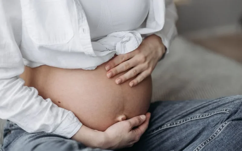 Close-up of a pregnant woman sitting, cradling her bare belly with both hands.
