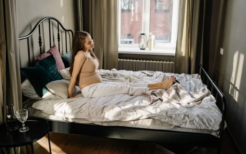 A pregnant woman sitting on a bed in a comfortable, sunlit bedroom.