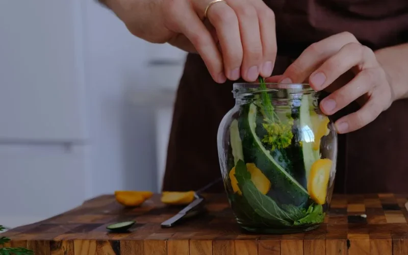 Hands organizing fresh vegetables for pickling in a glass jar on a wooden countertop.