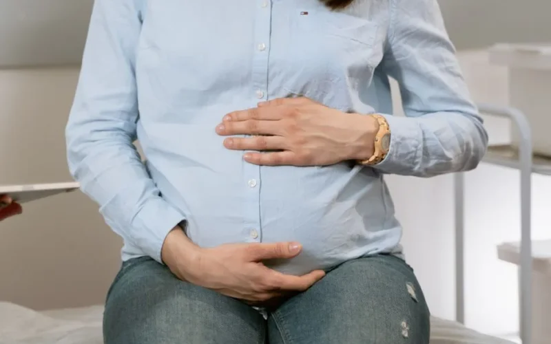 Pregnant woman sitting indoors, hands on belly, wearing casual clothes. Healthcare consultation setting.