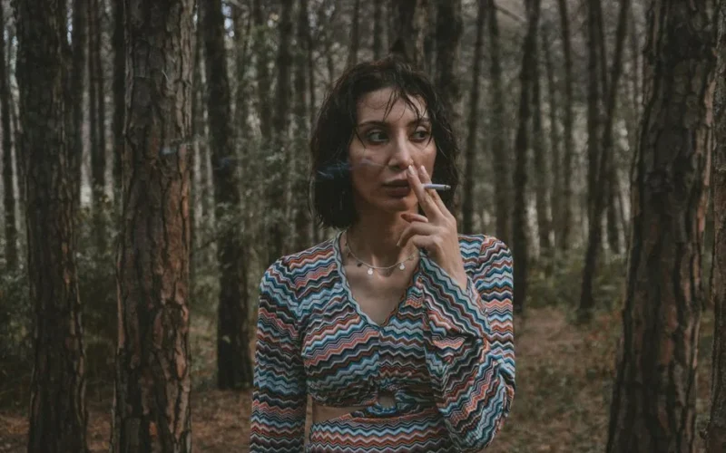 Woman standing in forest smoking a cigarette with pensive expression.