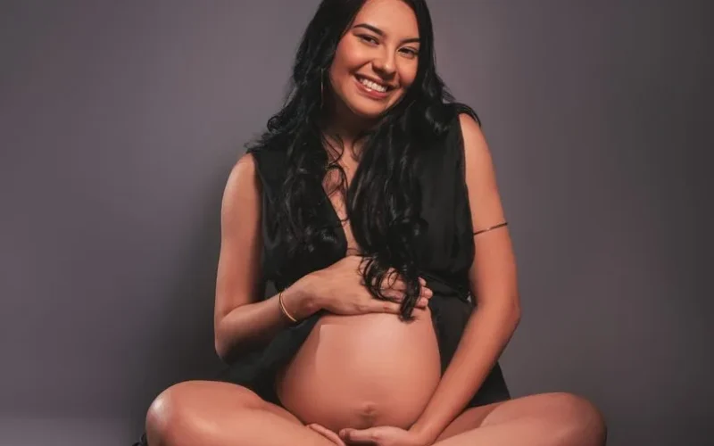 Beautiful pregnant woman smiling, seated in a studio with a gray background.
