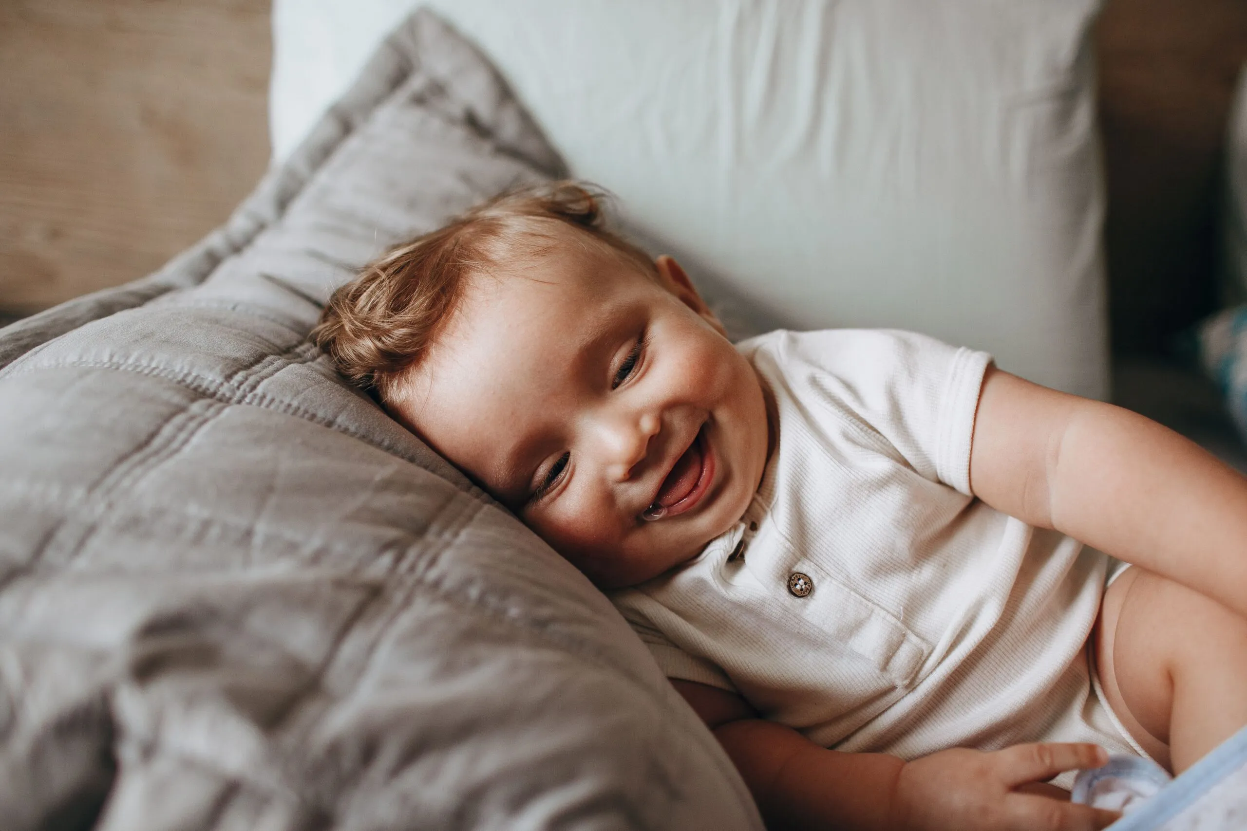 A cute baby in a white romper smiles happily while lying on a soft pillow.
