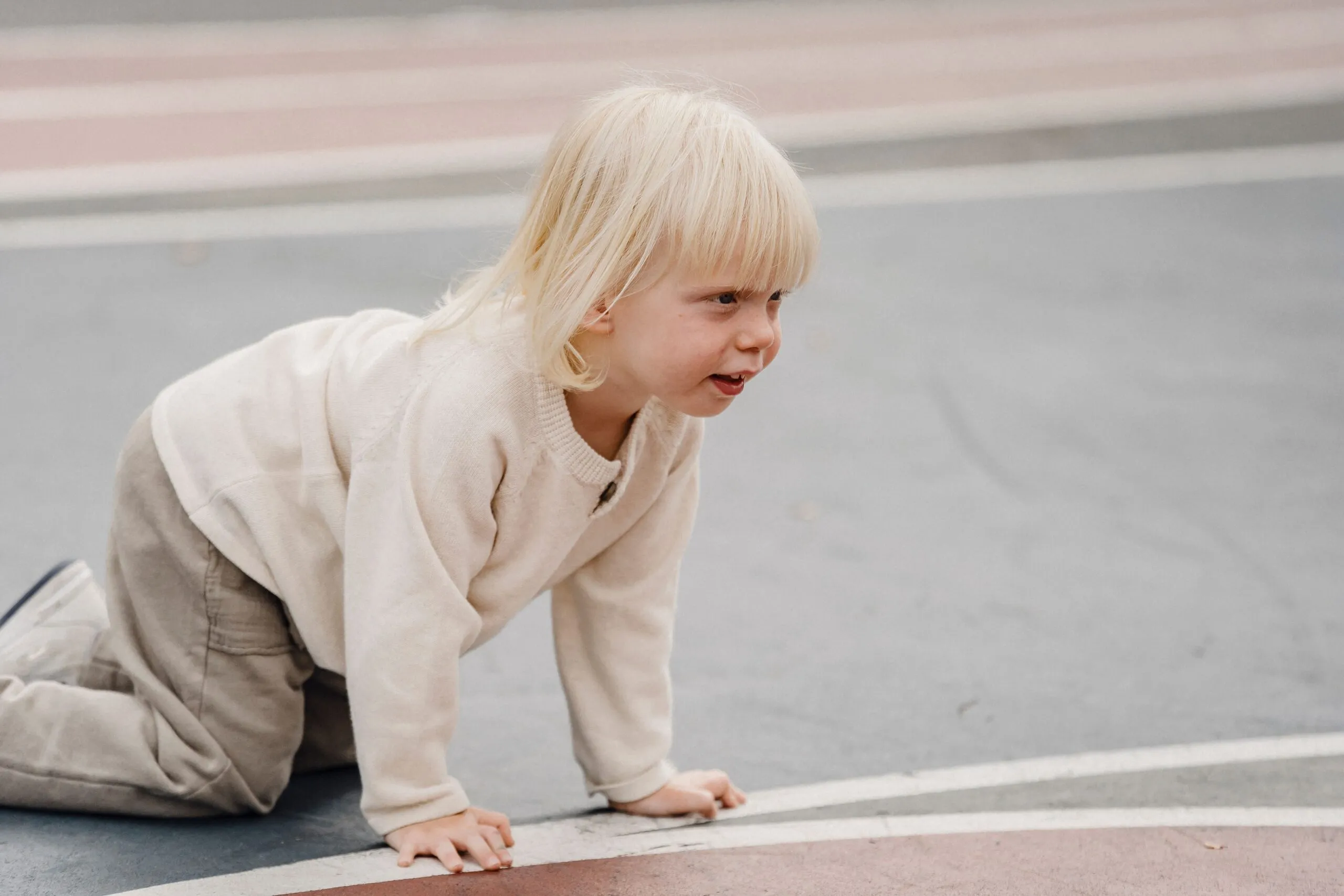 Side view of adorable cheerful little kid with blond hair standing on all fours on sports ground against blurred background