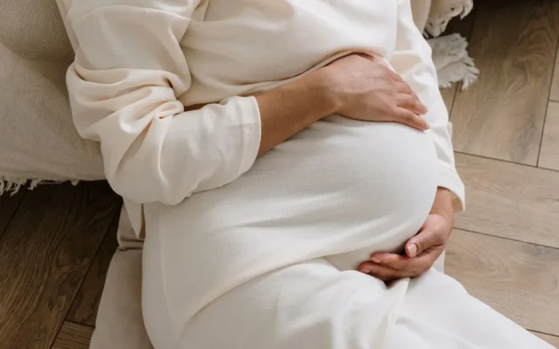 A serene moment of a pregnant woman in a cozy white dress holding her belly.