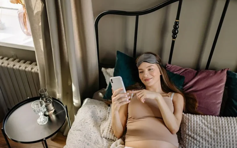 Pregnant woman lying in bed with cellphone, enjoying a relaxing morning indoors.