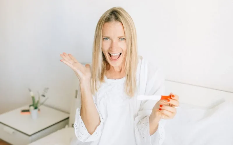 A joyful woman celebrates a positive pregnancy test while sitting on her bed.