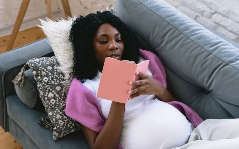 From above of focused pregnant African American female taking notes in notepad while resting on comfortable sofa in living room