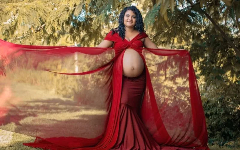 Pregnant woman in a flowing red dress posing confidently outdoors, showcasing her baby bump.