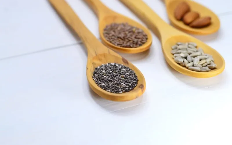 A variety of seeds and nuts in wooden spoons displayed on a white surface.