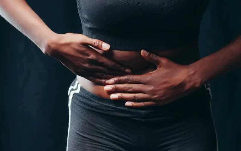 Close-up of a person pressing hands on abdomen, indicating discomfort or pain, against a black background.