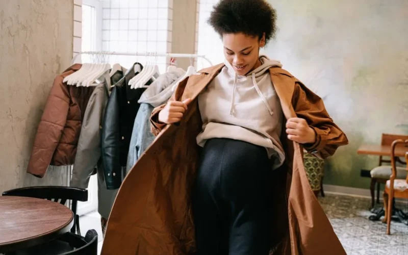 Pregnant woman in stylish coat adjusting outfit indoors near clothes rack.