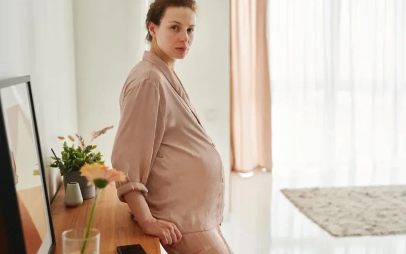 A pregnant woman wearing sleepwear leans on a wooden drawer in a bright, serene room.