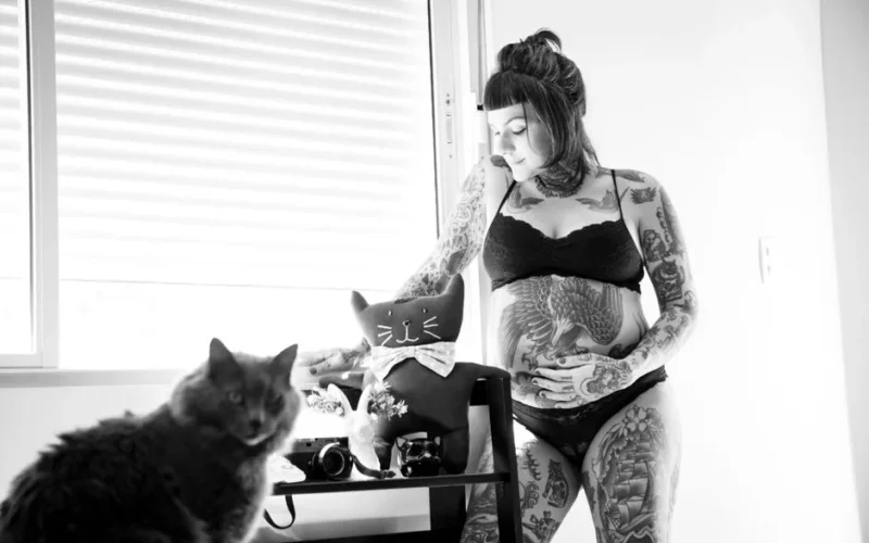 Intriguing black and white photo of a tattooed woman with a baby bump indoors with a cat.