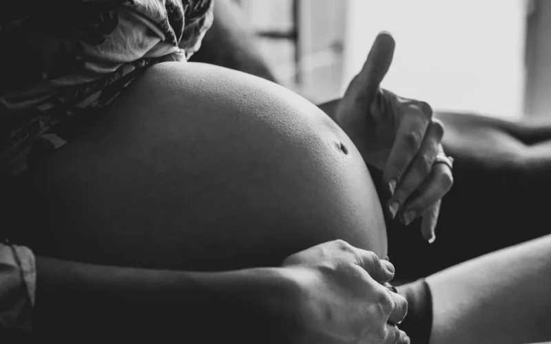 Side view of crop anonymous pregnant lady in crop top and panties sitting on sofa and caressing tummy