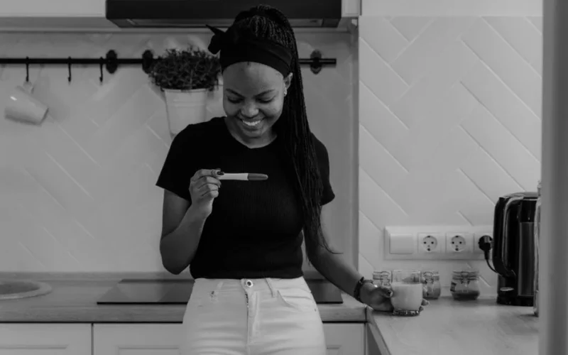 A smiling woman in a kitchen holds a pregnancy test, expressing happiness and excitement.
