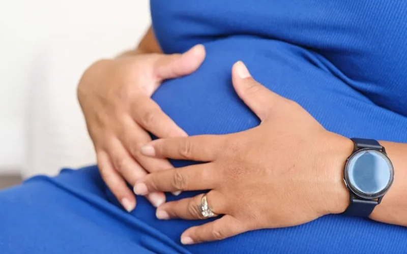 Closeup of a pregnant woman resting her hands on her belly, displaying a watch and a ring.