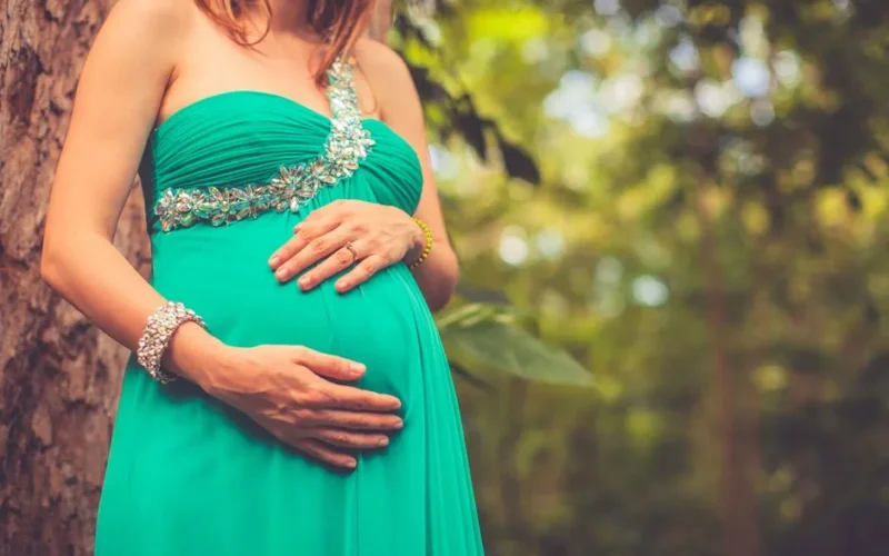 A pregnant woman wearing a green dress gently holds her belly in a serene outdoor setting.
