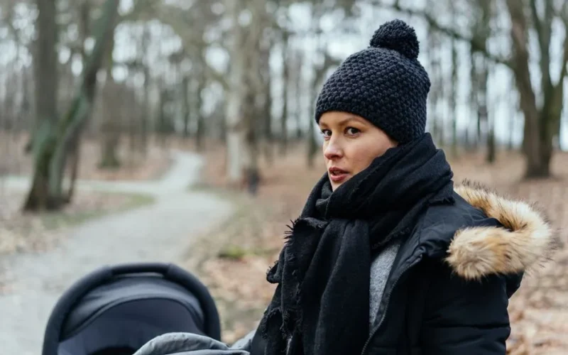 A woman in a beanie and scarf walking with a stroller through a park on a winter day.