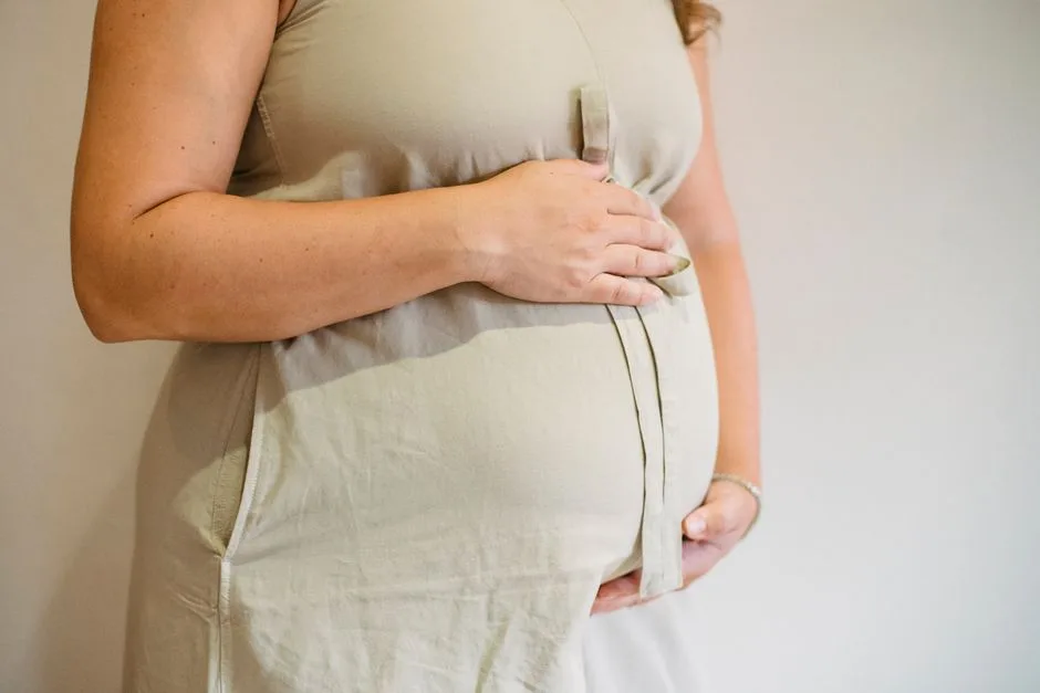 Crop faceless pregnant woman in casual dress standing near white wall and holding belly with hands