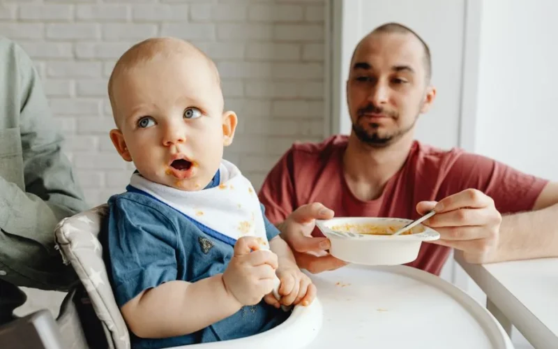 A father lovingly feeds his baby in a high chair, showcasing a family bonding moment.