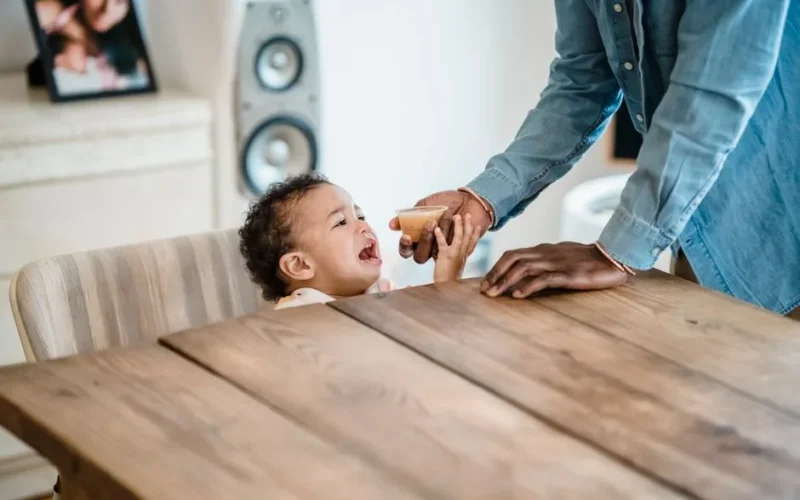 A father feeds his crying toddler applesauce at a wooden table indoors.