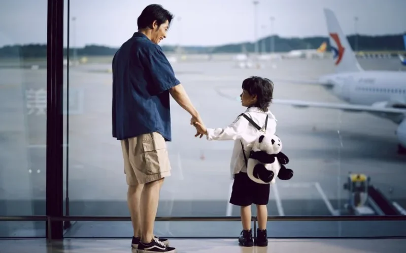 A father and child holding hands, gazing at planes through an airport window.
