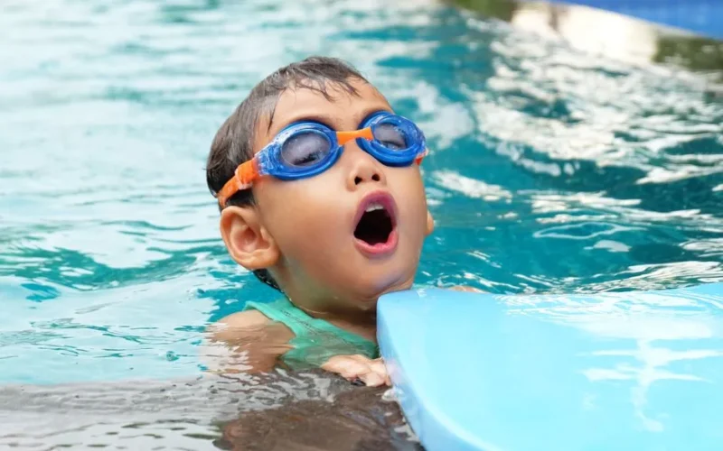 Young child swimming in a pool wearing blue goggles, enjoying summer.