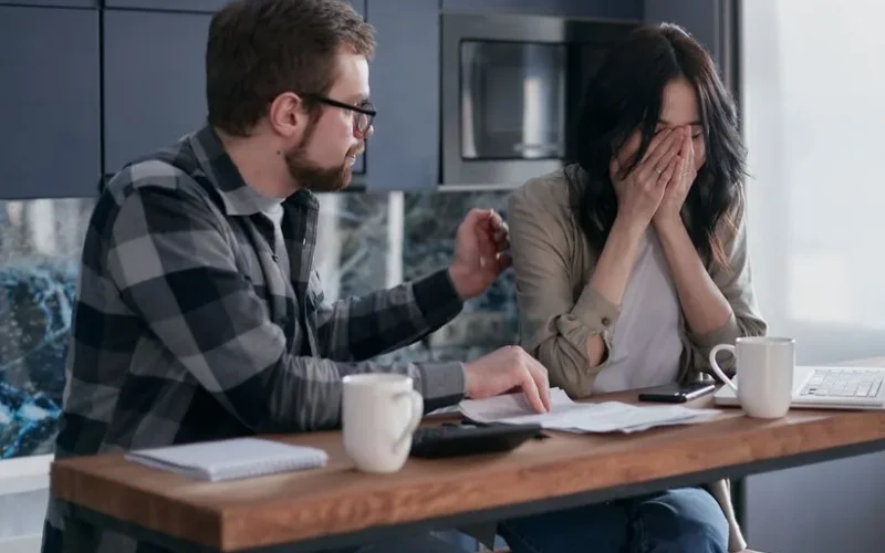 A couple discusses financial issues with bills and a laptop on the table, inside a modern kitchen setting.