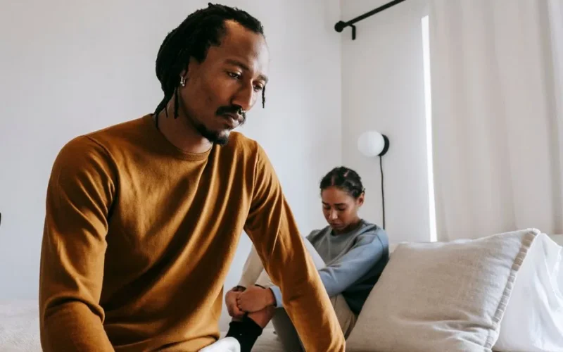 Young sad African American male near frustrated female partner sitting on bed during conflict at home