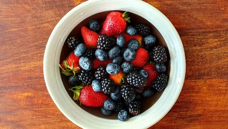 A vibrant assortment of strawberries, blackberries, and blueberries in a bowl on a wooden surface.