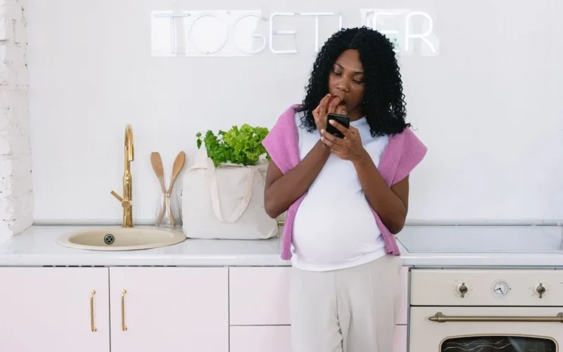 Focused pregnant African American female eating healthy food and browsing cellphone while standing in light kitchen with signboard at home
