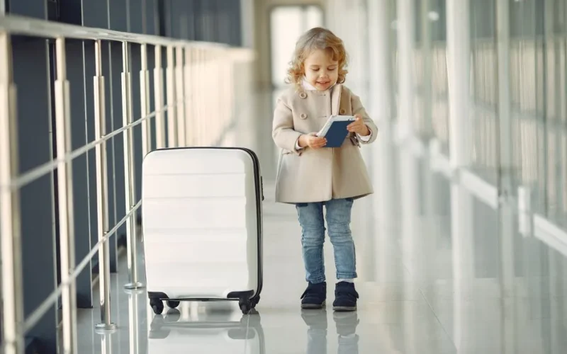 Full body of smiling cute little girl in jeans and beige coat standing near suitcase and checking information in documents