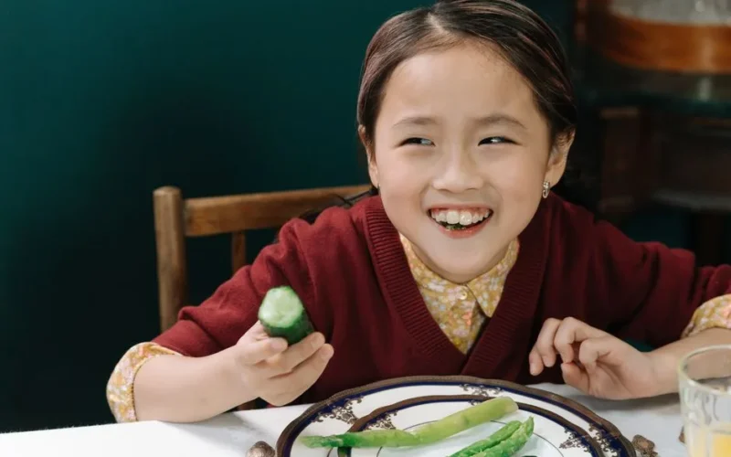 A happy child in a maroon sweater enjoying a meal with fresh vegetables at a dining table.