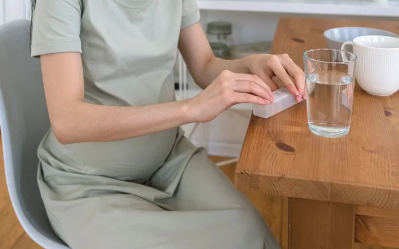 Unrecognizable pregnant female sitting at wooden table with glass of water while taking prenatal vitamins in light kitchen at home