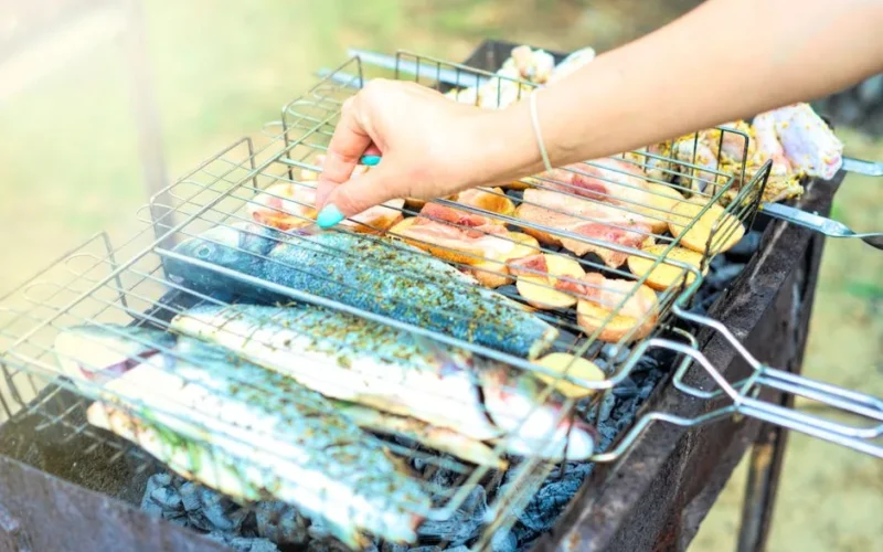 Outdoor barbecue grilling fish and meat. Woman's hand adjusting grill on a sunny summer day.