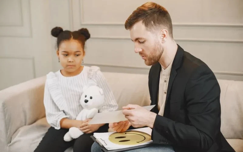 A child holding a plush toy listens to a man in a therapy session.