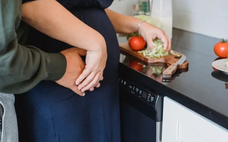 Side view of crop anonymous husband embarrassing pregnant wife preparing healthy salad in kitchen at home