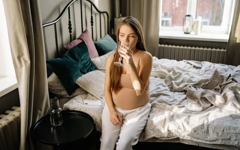 Pregnant woman sitting on bed, drinking water in a cozy bedroom setting.