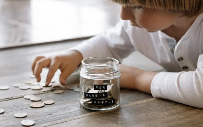 A young child collects coins in a jar labeled 'For Barbie Castle', symbolizing saving and dreams.