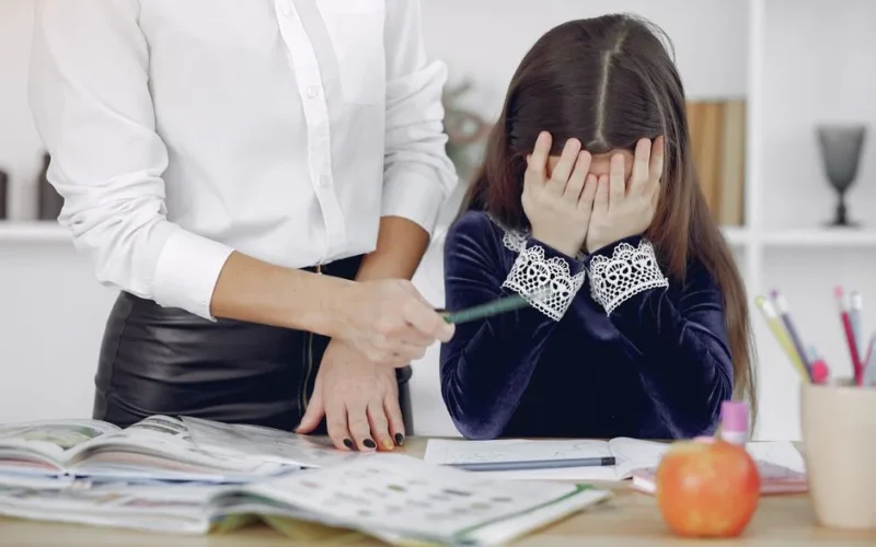 Little schoolgirl covering face with hands and crying while unrecognizable woman persuading to do homework and writing in copybook in modern workplace