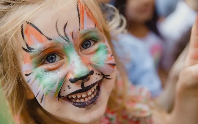 A cheerful young girl with vibrant animal face paint smiles brightly at an outdoor event.