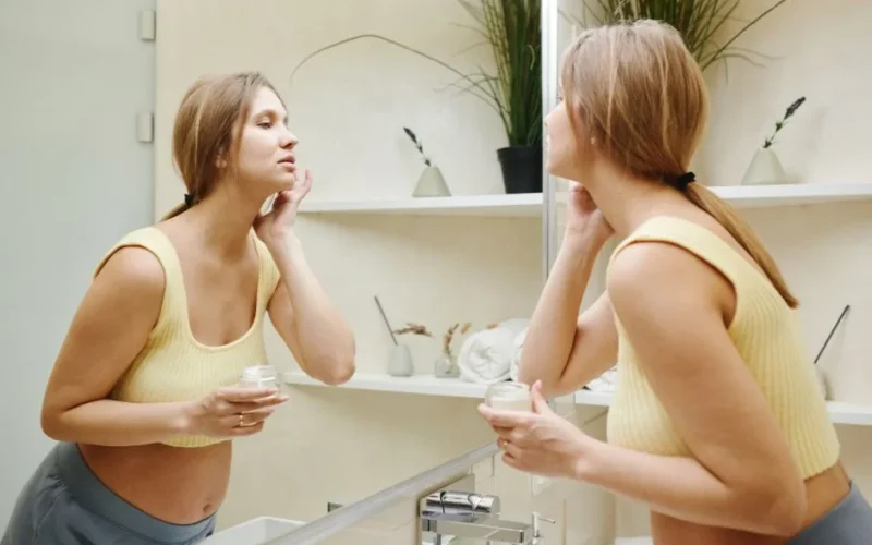 A pregnant woman gently applies skincare cream while looking into a bathroom mirror, promoting maternity care.