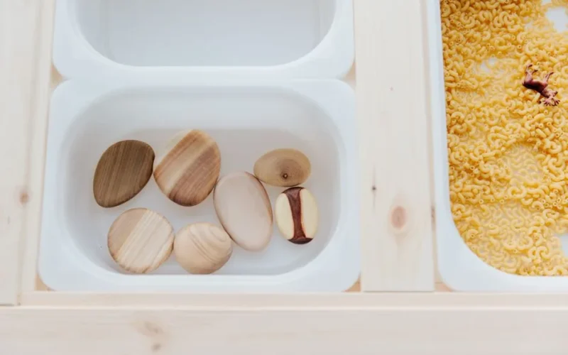 Set of various white plastic containers with stones and raw pasta for child sensory development on wooden table in light room